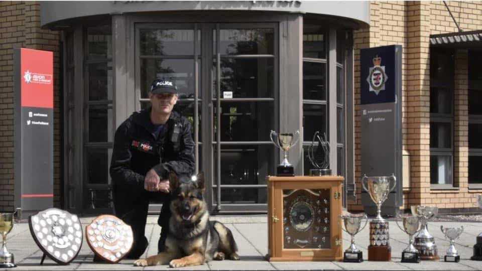 Handler and dog with a display of competition trophies and shields outside the police station